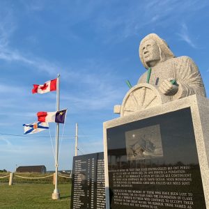 Monument en mémoire des pêcheurs perdus en mer, avec en arrière-plan les drapeaux de la Nouvelle-Écosse, de l'Acadie et du Canada qui flottent au vent